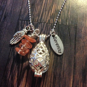 May include: Silver-toned necklace featuring a filigree teardrop pendant, orange gemstone chips, a charm with the word "Serenity", and a hand with wings charm. The necklace hangs on a ball chain.
