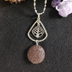 May include: Silver-toned necklace featuring a pendant with a tree design within a diamond shape, and a round, brown lava stone. The necklace is displayed on a black surface, with crystals in the background.