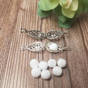 May include: Two silver-colored, filigree-style essential oil diffuser pendants, one open and one closed with a white cotton ball inside. Several white cotton balls are scattered below. The background is a wooden surface.
