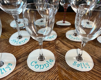 May include: A set of six white wine glass coasters with blue embroidered names: Arlene, Kathy, Mary, Lisa, Connie, and Sally. The coasters are on a wooden table with six wine glasses.
