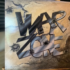 Op de afbeelding: Een vintage vinyl platenhoes met een verweerde, textuurrijke achtergrond. De albumtitel "WAR ZONE" is geschreven in vetgedrukte, zwarte letters met een metallic glans. De letters zijn gelaagd en overlappen elkaar, wat een dynamisch en edgy ontwerp creëert. De hoes heeft verschillende kogelgaten, wat bijdraagt aan de rauwe en intense esthetiek.