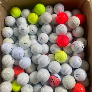 Könnte beinhalten: Ein Karton voller Golfbälle. Die Golfbälle sind überwiegend weiß, mit einigen gelben und roten Bällen. Die Golfbälle tragen verschiedene Markennamen und Nummern.