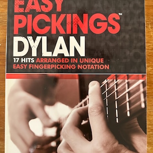 May include: A book cover with the title "EASY PICKINGS DYLAN" in red and white text. The cover features a photo of hands playing a guitar. The text below reads "17 HITS ARRANGED IN UNIQUE EASY FINGERPICKING NOTATION."