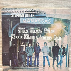 May include: Album cover for Stephen Stills' band Manassas. The cover features a group of seven people standing in front of a train station. The album title and band members' names are printed at the top of the cover.