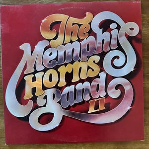 Könnte beinhalten: Ein Vintage-Vinyl-Albumcover mit dem Text "The Memphis Horns Band II" in einer Retro-Schriftart. Das Albumcover ist rot mit dem Text in Orange-, Weiß- und Silbertönen.