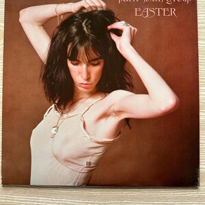 Puede incluir: Portada del álbum "Easter" de Patti Smith Group. La portada presenta a una mujer con cabello oscuro, vistiendo una blusa de color claro, posando con los brazos levantados. El título del álbum y el nombre del artista están en una fuente estilizada sobre un fondo marrón.