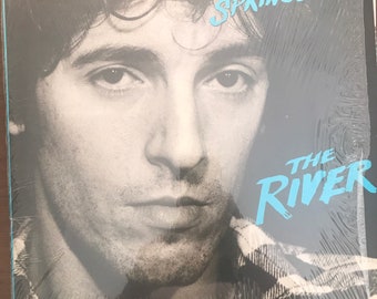 May include: A vinyl record album cover featuring a black and white portrait of Bruce Springsteen wearing a plaid shirt. The text BRUCE SPRINGSTEEN appears in light blue at the top right, and the text THE RIVER appears in light blue at the bottom right.