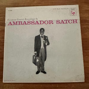 May include: Vintage vinyl record album cover for "Ambassador Satch" by Columbia Records. The cover features a black and white image of a man in a suit holding a briefcase and a trumpet. The album title is in pink lettering.