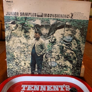 Könnte beinhalten: Ein Vintage-Vinyl-Plattencover mit einem Mann in einer Waldlandschaft. Der Albumtitel lautet "Junior Samples Moonshining" und das Plattenlabel ist "Pickwick 33".