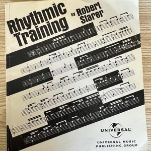 May include: A vintage music book titled "Rhythmic Training" by Robert Starer. The cover features black musical notes and staff lines on a cream background. The Universal Music Publishing Group logo is visible.