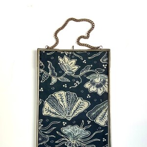 May include: A framed piece of batik fabric with a white floral pattern on a dark blue background. The frame is silver and has a chain for hanging.