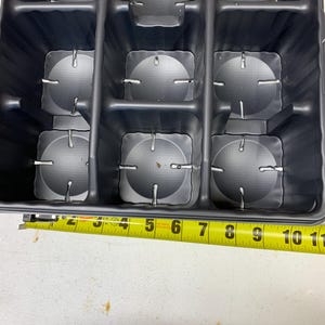 May include: Black plastic tray with nine circular compartments, each with three small holes. A yellow measuring tape is placed below the tray, showing measurements from 2 to 11 inches.