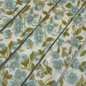 Puede incluir: Primer plano de tela con estampado floral. La tela tiene un fondo blanco con un patrón repetido de flores azules y hojas verdes. La tela está cortada en tiras.