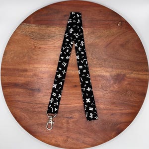 May include: Black lanyard with white mathematical symbols, including numbers, plus signs, and division symbols. The lanyard has a silver-colored metal clasp at the bottom. The lanyard is displayed on a wooden surface.