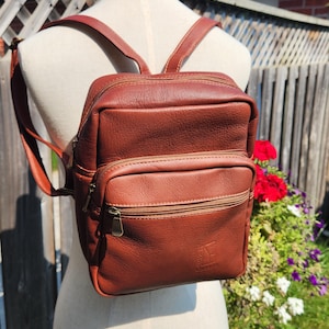 Puede incluir: Una mochila de cuero marrón medio con un compartimento principal con cremallera y un bolsillo frontal más pequeño con cremallera. La bolsa tiene correas ajustables para los hombros y un pequeño logotipo en relieve en la esquina inferior derecha.
