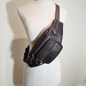 Puede incluir: Un bolso de cintura de cuero marrón oscuro con múltiples compartimentos con cremallera y una correa marrón ajustable. El bolso, con cremalleras y costuras doradas, se exhibe en un maniquí.