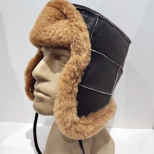 Peut inclure: Un bonnet d'aviateur en peau de mouton marron avec des panneaux en cuir noir et une longue ficelle noire.