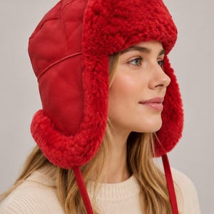 May include: A bright red trapper hat featuring a smooth leather exterior with quilted stitching and a thick, plush red shearling lining. The hat includes ear flaps that extend downwards with matching red leather ties hanging from the sides.