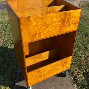 May include: A wooden storage cart with a warm, amber finish. The cart has multiple compartments and sits on four caster wheels. The design features a sloped top and a series of shelves, suitable for organizing items.