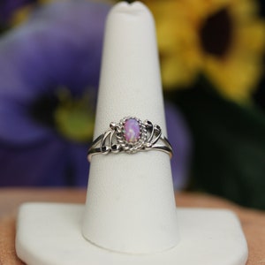 May include: A silver ring with a pink opal gemstone set in a delicate, ornate design. The ring is displayed on a white ring stand.