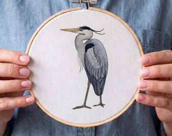 Pode incluir: Um bastidor de bordado circular de madeira contendo tecido branco com uma ilustração detalhada de linha multicolorida de uma garça-real em pé de perfil. A ave apresenta tons de cinza, preto e amarelo, capturados em bordado fino.