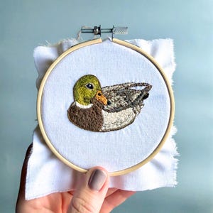 Puede incluir: Bordado a mano de un pato en un bastidor de madera. El pato tiene la cabeza verde, el cuerpo marr&oacute;n y las alas grises. El bordado est&aacute; sobre tela blanca con un borde sin rematar. El bastidor es sostenido por una mano.