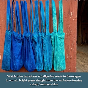 May include: A row of indigo-dyed tote bags in varying shades of blue and teal hang against a weathered red wooden background. The bags have long straps and are made of fabric. The text on the image reads: "Watch color transform as indigo dye reacts to the oxygen in our air, bright green straight from the vat before turning a deep, luminous blue."