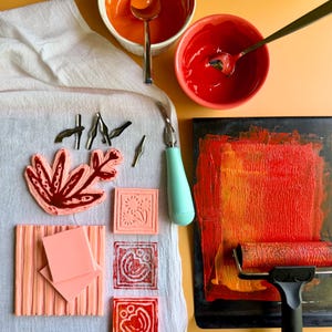 May include: An overhead shot of art supplies, including bowls of orange and red paint, a paint roller, carving tools, and various carved stamps. The stamps feature floral and geometric designs. A light blue handled carving tool is also present.
