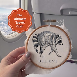 May include: A circular wooden embroidery hoop containing white fabric with a black and white stitched illustration of a raccoon. The word BELIEVE is embroidered in black capital letters below the raccoon. A red graphic overlay says The Ultimate Travel Craft.