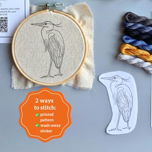Pu&ograve; includere: Un kit da ricamo con un telaio in legno contenente un motivo a linee di un airone su tessuto beige, un adesivo idrosolubile con un airone e fili da ricamo in grigio, blu, giallo e bianco. Include il testo: 2 ways to stitch.