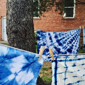 May include: Several pieces of blue and white tie-dye fabric hang on a clothesline, secured with wooden clothespins. The fabrics display various patterns, including spiral and geometric designs. A brick building and a tree are in the background.