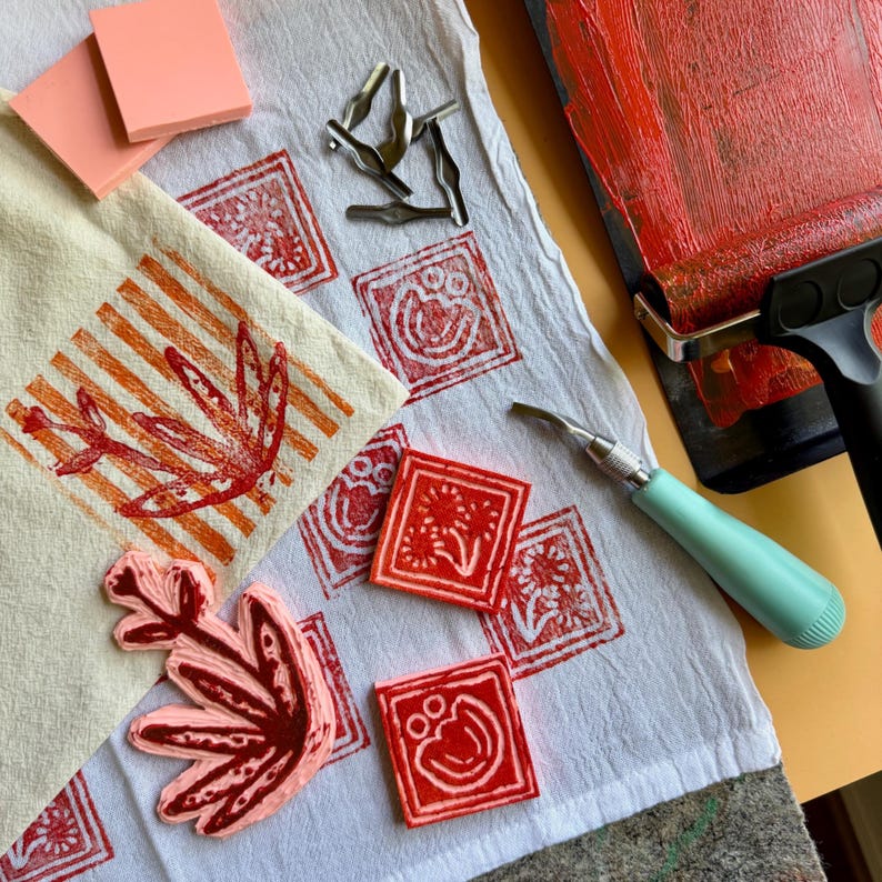 May include: A collection of linocut printmaking supplies. Includes red ink, a brayer, carving tools, and carved linoleum blocks with floral and geometric designs. A printed linen towel with orange leaf and stripe patterns is also present.