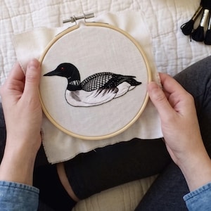 May include: A circular wooden embroidery hoop containing a piece of white fabric with a detailed thread-stitched design of a loon. The bird features a black head with a red eye, a white neck band, and black and white patterned wing feathers.