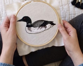 May include: A circular wooden embroidery hoop containing a piece of white fabric with a detailed thread-stitched design of a loon. The bird features a black head with a red eye, a white neck band, and black and white patterned wing feathers.