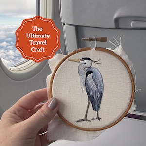 Pu&ograve; includere: Un telaio da ricamo contenente un pannello in tessuto con un'illustrazione dettagliata cucita a filo di un airone cenerino. L'uccello presenta sfumature di filo blu, grigio e nero. Un grafico arancione smerlato include il testo The Ultimate Travel Craft.