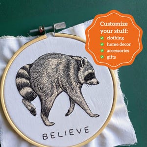 May include: A round wooden embroidery hoop containing white fabric with a detailed black and white thread-stitched raccoon and the word BELIEVE. An orange graphic overlay reads: Customize your stuff: clothing, home decor, accessories, gifts.