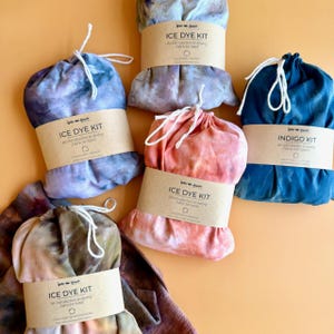 Pode incluir: Uma variedade de kits de tingimento com gelo em v&aacute;rias cores, incluindo azul, rosa e roxo. Cada kit &eacute; embalado em um saco com cord&atilde;o e embrulhado com uma faixa de papel castanho que diz "ICE DYE KIT" e "An introduction to dyeing fabric by hand."