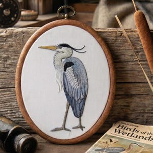 May include: An oval embroidery hoop containing a white fabric panel with a detailed thread-stitched illustration of a grey heron. The bird features shades of blue, grey, and white thread, a long yellow beak, and a dark crest, framed in wood.