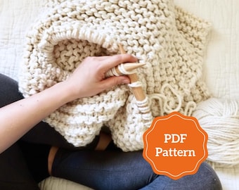 May include: Cream-colored, chunky knit blanket with wooden knitting needles. A ball of yarn sits nearby. The blanket is being knitted, with the text "PDF Pattern" on an orange badge.