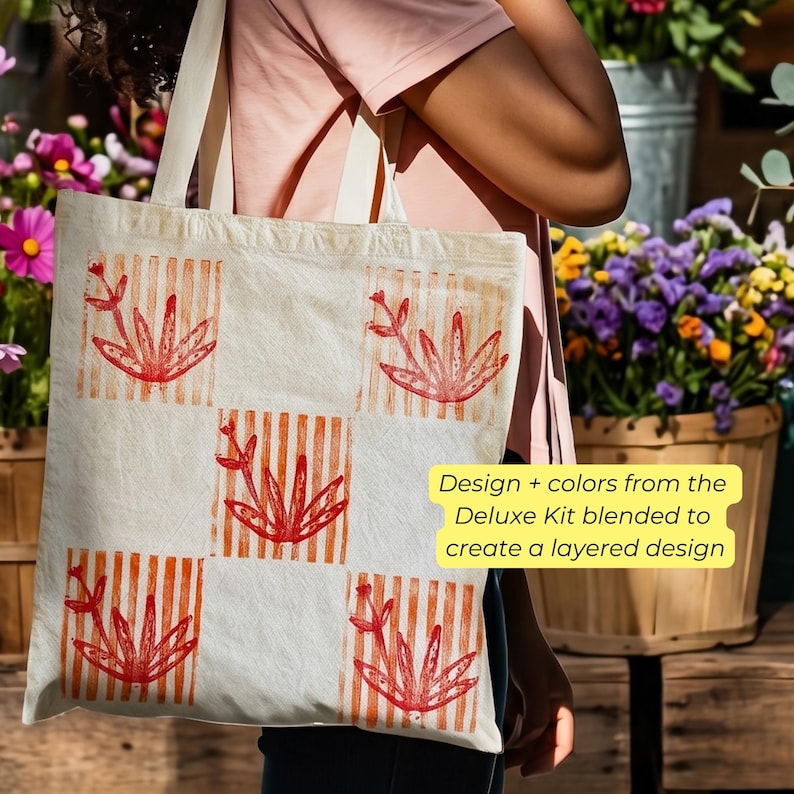 May include: Cream-colored tote bag with a repeating pattern of red floral designs and orange striped squares. The bag is carried by a person. Text on the bag reads: Design + colors from the Deluxe Kit blended to create a layered design.