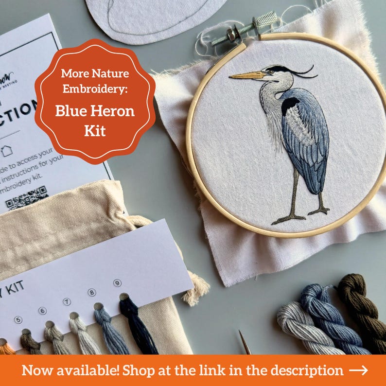 May include: An embroidery kit featuring a wooden hoop with a blue heron design stitched on white fabric. The kit includes a thread organizer card with numbered slots, various shades of embroidery floss, and an instruction card with a QR code.