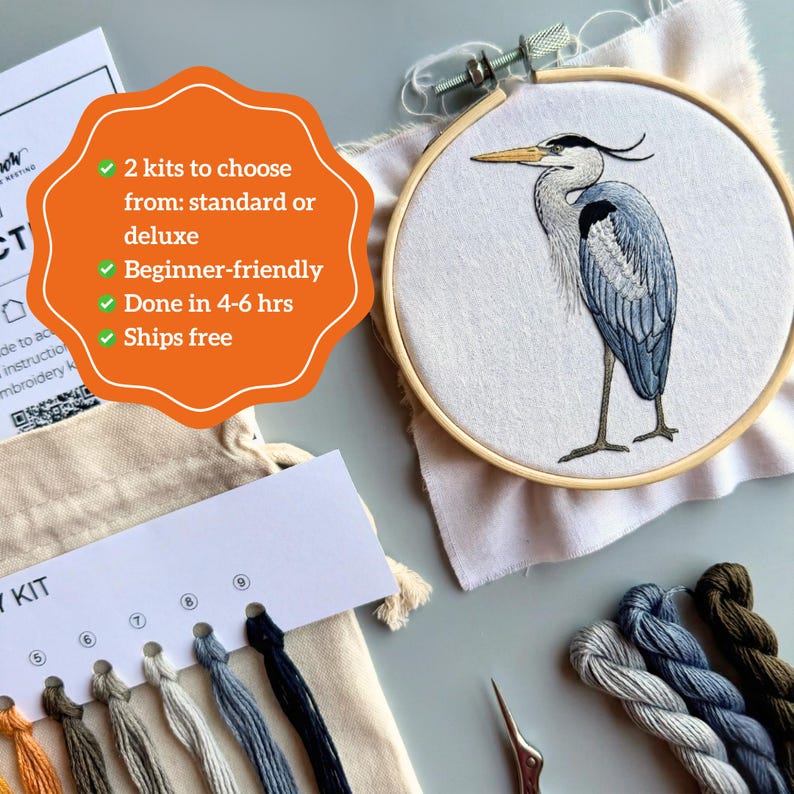 May include: An embroidery kit featuring a circular wooden hoop with a white fabric panel embroidered with a grey and blue heron. The kit includes a thread organizer card with numbered holes, various colored embroidery floss, and a small metal tool.