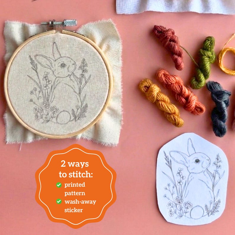 May include: An embroidery hoop containing fabric with a printed rabbit and floral pattern, accompanied by several small skeins of colored embroidery thread and a separate paper sticker featuring the same rabbit design with text 2 ways to stitch.