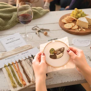 Puede incluir: Kit de bordado con un dise&ntilde;o de pato. El kit incluye un bastidor de madera, hilos de varios colores, instrucciones y un patr&oacute;n. Una copa de vino tinto, queso y galletas tambi&eacute;n est&aacute;n sobre la mesa.