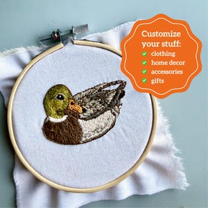 Puede incluir: Un pato bordado en un aro de madera. El pato presenta puntadas detalladas en tonos marrones, verdes y blancos. El texto "Customize your stuff: clothing, home decor, accessories, gifts" est&aacute; en una forma naranja.