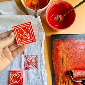 May include: A square, red stamp with a white floral design is held over a white cloth. Several other stamped images are visible on the cloth. Bowls of orange and red paint and a paint roller are also in the image, suggesting a DIY craft project.