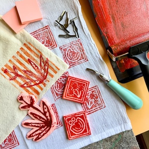 May include: An overhead shot of printmaking supplies. A white cloth displays red block prints and a leaf design. Pink foam blocks and carving tools are scattered around. A red ink roller and a carving tool with a blue handle are also present.