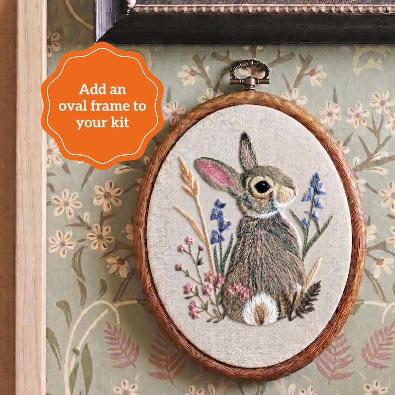 May include: An oval embroidery hoop containing a fabric panel with a detailed thread-stitched rabbit surrounded by blue, pink, and wheat-colored floral elements. The text 'Add an oval frame to your kit' is displayed in an orange scalloped graphic.