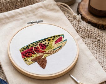 May include: A circular wooden embroidery hoop containing white fabric with a detailed thread-stitched design of a trout fish head. The fish features vibrant green, yellow, and red scales with black spots and an open mouth, rendered in embroidery.