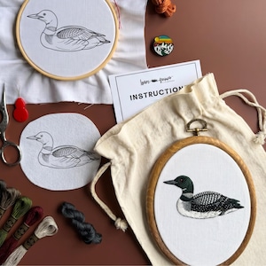 May include: An embroidery kit featuring a wooden hoop with a finished loon design, a second hoop with a printed loon outline, a fabric swatch with a loon outline, an instruction card, a small enamel pin, embroidery floss, and a drawstring bag.